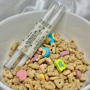 May include: Two cuticle oil pens with the text "Lucky you CUTICLE OIL" rest on a bowl of cereal with colorful marshmallows. The pens have a clear tube with a white cap and a label with pastel designs.