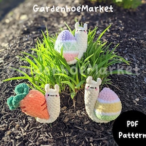 May include: Three crocheted snail amigurumi toys with different colored shells. One snail has a green and orange carrot shell, one has a purple, green, and pink Easter egg shell, and one has a pink, yellow, and green Easter egg shell. The snails are sitting on a bed of green grass and brown mulch. The text "GardenhoeMarket" is visible at the top of the image. The text "PDF Pattern" is visible at the bottom of the image.