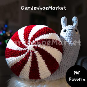 May include: Crochet pattern for a peppermint candy snail amigurumi. The snail is made with red and white yarn and has a gray body. The pattern is available as a PDF download.
