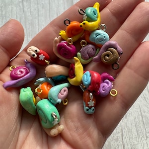 Puede incluir: Una colección de encantos de caracol de arcilla polimérica hechos a mano, de colores vivos, con anillos de salto de plata. Los caracoles son de varios colores, incluyendo verde, azul, amarillo, naranja, rojo y morado. Algunos caracoles tienen flores blancas en sus conchas.