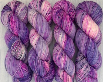 May include: Four skeins of hand-dyed yarn in shades of purple, pink, and white. The yarn is twisted into a skein shape, with visible color variations and speckles. The yarn is arranged on a light wood surface.