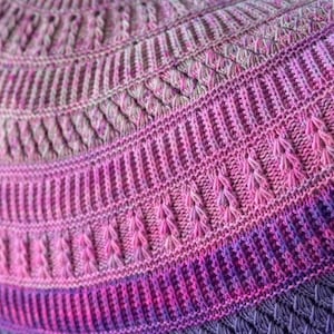 Könnte beinhalten: Nahaufnahme einer gestrickten Decke mit einem Farbverlauf von Rosa und Lila. Die Decke hat ein strukturiertes Muster mit verschiedenen Maschen.