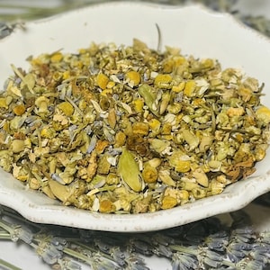 May include: A white ceramic bowl filled with a blend of dried herbs, including chamomile, lavender, and other botanicals. The herbs are a light green and yellow color.