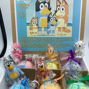 May include: A white box with a blue and white label featuring the cartoon characters Bluey, Bingo, and Bandit from the show Bluey. The box contains six bath bombs with Bluey character toys inside. The bath bombs are wrapped in clear plastic and have a variety of colors and scents. The label on the box says "Bluey Bath Bomb Gift Set" and "Six (6) Surprise Bluey Toys Inside!"