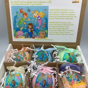 May include: A gift set of six mermaid-themed bath bombs, each 5.5 oz, in a cardboard box. The box includes bath bombs in various colors and scents, each wrapped in cellophane and tied with a ribbon. The box also features a mermaid illustration.