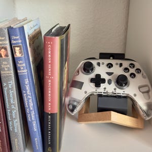May include: A clear and white video game controller rests on a wooden stand, next to a row of books with titles like "Dear America" and "Common Sense Uncommon Man." The controller has black buttons and a black top mount.