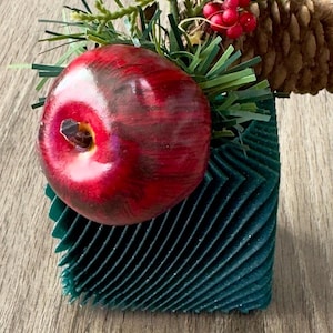 May include: A decorative arrangement featuring a large, glossy red apple, green pine needles, red berries, and a pine cone. The apple rests in a teal, textured, geometric holder, creating a festive, seasonal display.