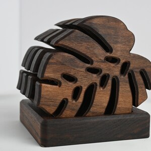 May include: A set of dark brown wooden coasters shaped like a monstera leaf. The coasters are stacked on top of each other and have a square base.