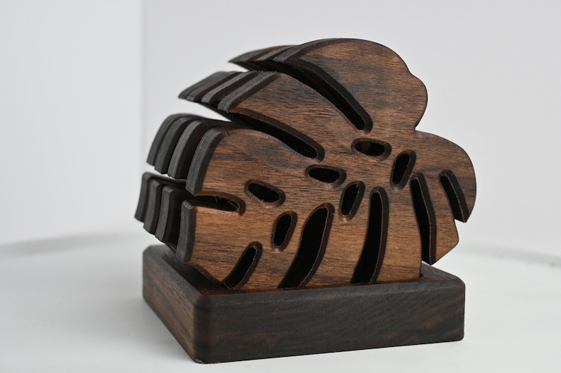 May include: A set of dark brown wooden coasters shaped like a monstera leaf. The coasters are stacked on top of each other and have a square base.
