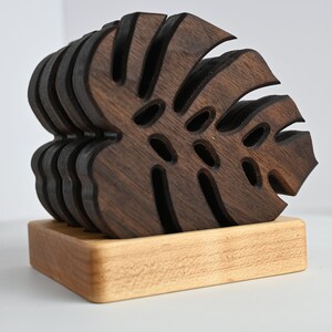 May include: A set of six dark brown wooden coasters shaped like monstera leaves, stacked on a light brown wooden base.