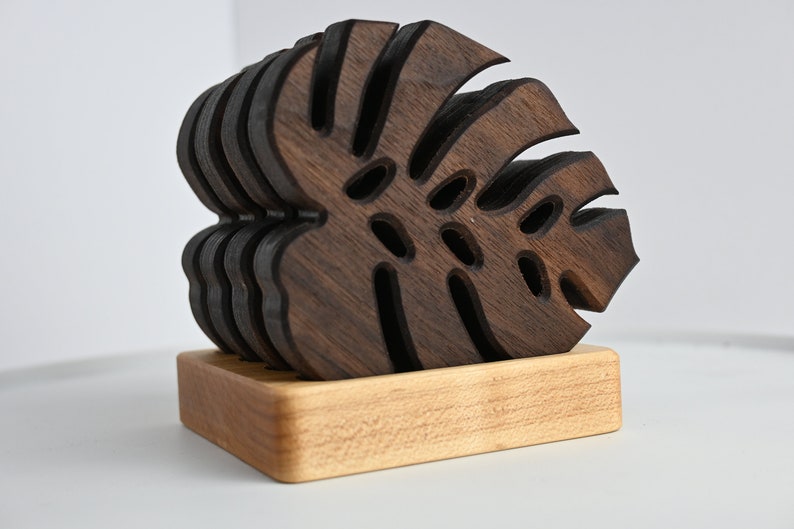 May include: A set of six dark brown wooden coasters shaped like monstera leaves, stacked on a light brown wooden base.