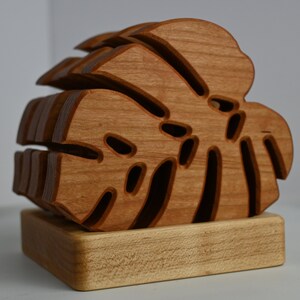 May include: A wooden coaster featuring a layered, intricately cut monstera leaf design. The coaster is made of natural wood and has a light brown color.
