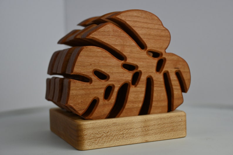 May include: A wooden coaster featuring a layered, intricately cut monstera leaf design. The coaster is made of natural wood and has a light brown color.