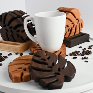 May include: Set of four wooden coasters featuring a tropical leaf design. The coasters are made of dark brown and light brown wood and are arranged on a white surface with coffee beans scattered around them. A white mug sits on top of one of the coasters.