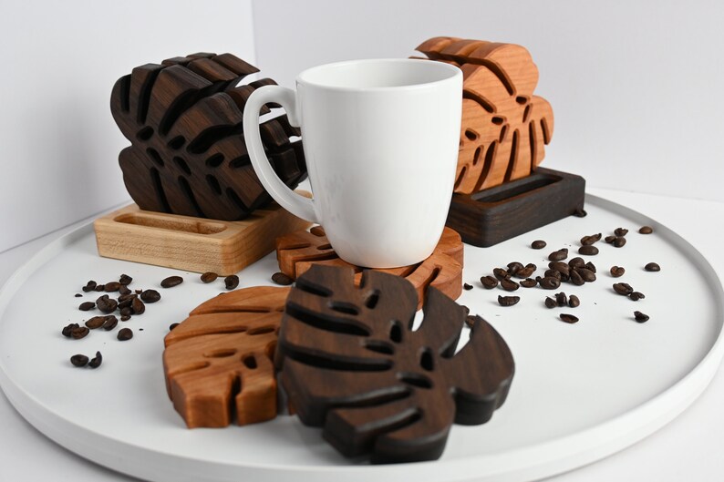 May include: Set of four wooden coasters featuring a tropical leaf design. The coasters are made of dark brown and light brown wood and are arranged on a white surface with coffee beans scattered around them. A white mug sits on top of one of the coasters.