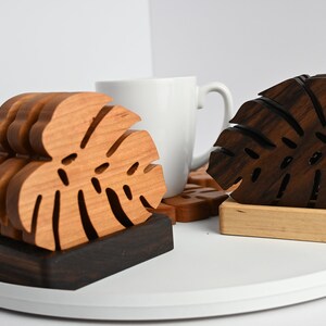 May include: Two sets of wooden coasters featuring a monstera leaf design. One set is made of light wood and the other is made of dark wood. The coasters are stacked on top of each other and are sitting on a white surface.
