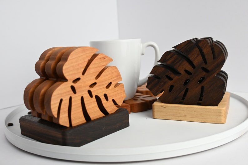 May include: Two sets of wooden coasters featuring a monstera leaf design. One set is made of light wood and the other is made of dark wood. The coasters are stacked on top of each other and are sitting on a white surface.