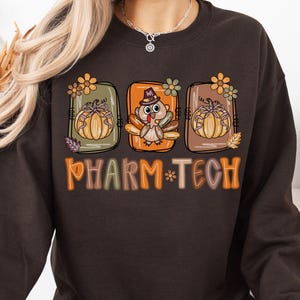 Peut inclure: Sweat-shirt chocolat foncé avec un graphisme coloré. Le graphisme présente trois motifs rectangulaires avec des citrouilles, des fleurs et une dinde portant un chapeau. Le texte "PHARM-TECH" est imprimé en orange.