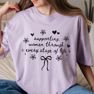 May include: A lavender t-shirt with a black graphic that reads "supporting women through every stage of life" with a bow and flowers.