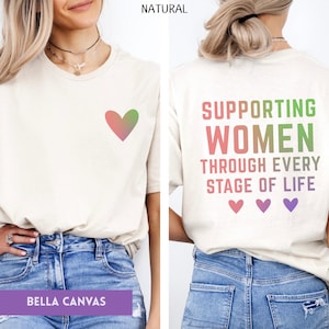 May include: Cream-colored t-shirt with a gradient heart graphic on the front. The back of the shirt features the text "SUPPORTING WOMEN THROUGH EVERY STAGE OF LIFE" in green, pink, and purple. Three heart graphics are below the text.