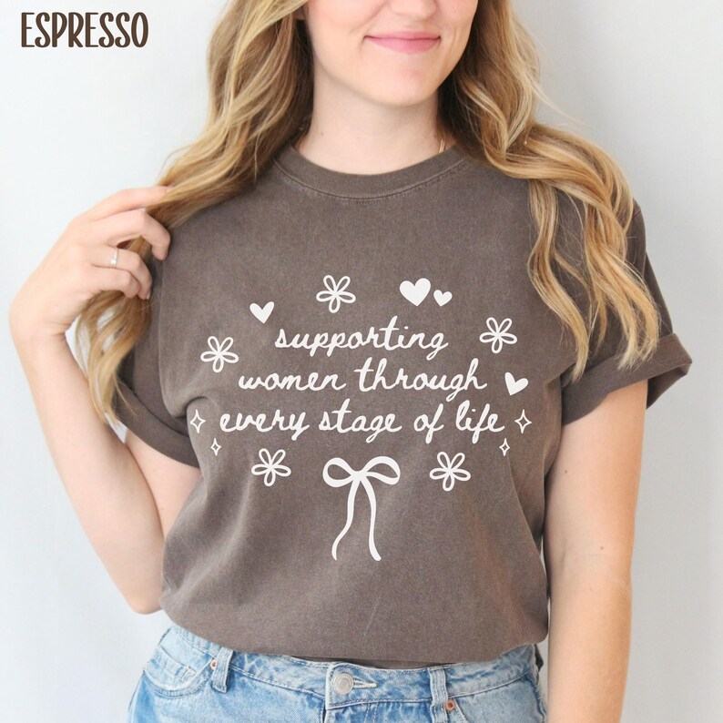 May include: A brown t-shirt with a white graphic that says "supporting women through every stage of life". The graphic also includes hearts, flowers, and a bow.