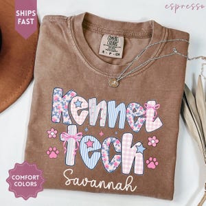 Könnte beinhalten: Ein braunes Comfort Colors T-Shirt mit dem Schriftzug "Kennel Tech Savannah" in einem farbenfrohen Design. Das Design umfasst Blumen-, Streifen- und Vichy-Muster sowie Pfotenabdrücke und Schleifen. Eine silberne Halskette liegt über dem Shirt.