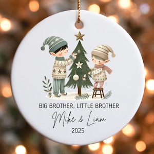 Könnte beinhalten: Runder, weißer Weihnachtsschmuck mit einer goldenen Aufhängeschlaufe. Er zeigt zwei Kinder, die einen Weihnachtsbaum schmücken. Der Text auf dem Ornament lautet "BIG BROTHER, LITTLE BROTHER", personalisierte Namen und das Jahr 2025.