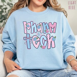 May include: Light blue sweatshirt with the words "Pharm Tech" in a colorful design. The letters are decorated with pink polka dots, blue stripes, and floral accents. Two pills are also featured.