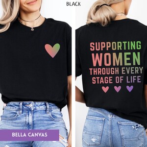 May include: Black t-shirt with a gradient heart graphic on the front. The back of the shirt features the text "SUPPORTING WOMEN THROUGH EVERY STAGE OF LIFE" with heart graphics. Made by Bella Canvas.