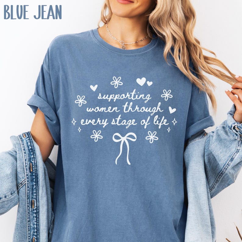 May include: A blue jean t-shirt with white text that reads "supporting women through every stage of life". The text is surrounded by small white hearts, stars, and flowers.
