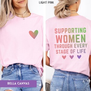 May include: Light pink Bella Canvas t-shirt with a heart graphic on the front and text on the back that reads "Supporting Women Through Every Stage of Life". The text and heart are in shades of pink and green.