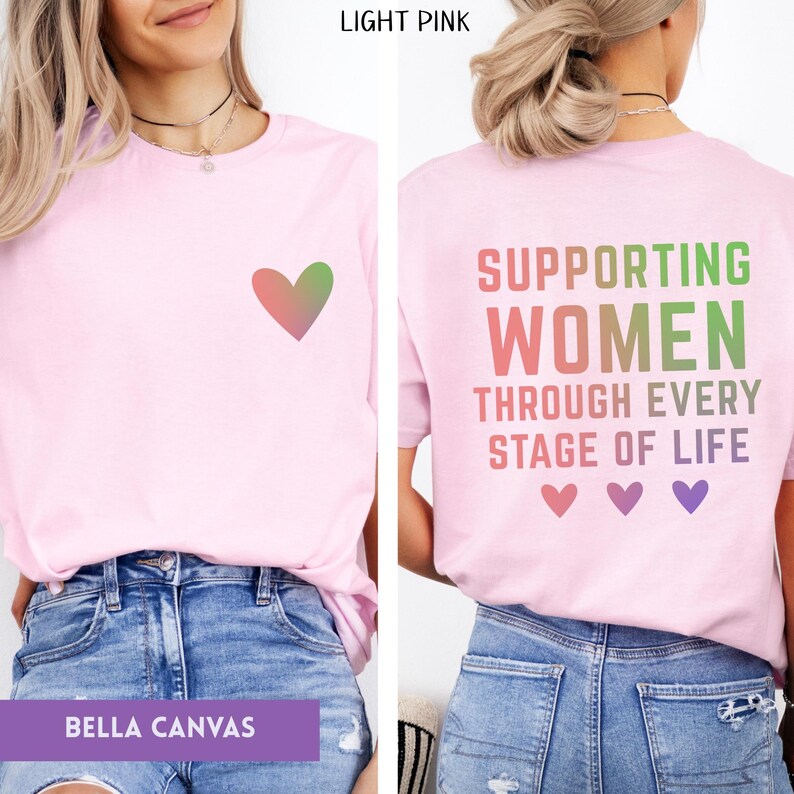 May include: Light pink Bella Canvas t-shirt with a heart graphic on the front and text on the back that reads "Supporting Women Through Every Stage of Life". The text and heart are in shades of pink and green.