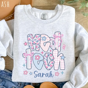 May include: A light gray sweatshirt with the words "Med Tech Sarah" in a colorful, floral, and striped design. The letters are decorated with pink, blue, and white patterns, including flowers, bows, and stars.