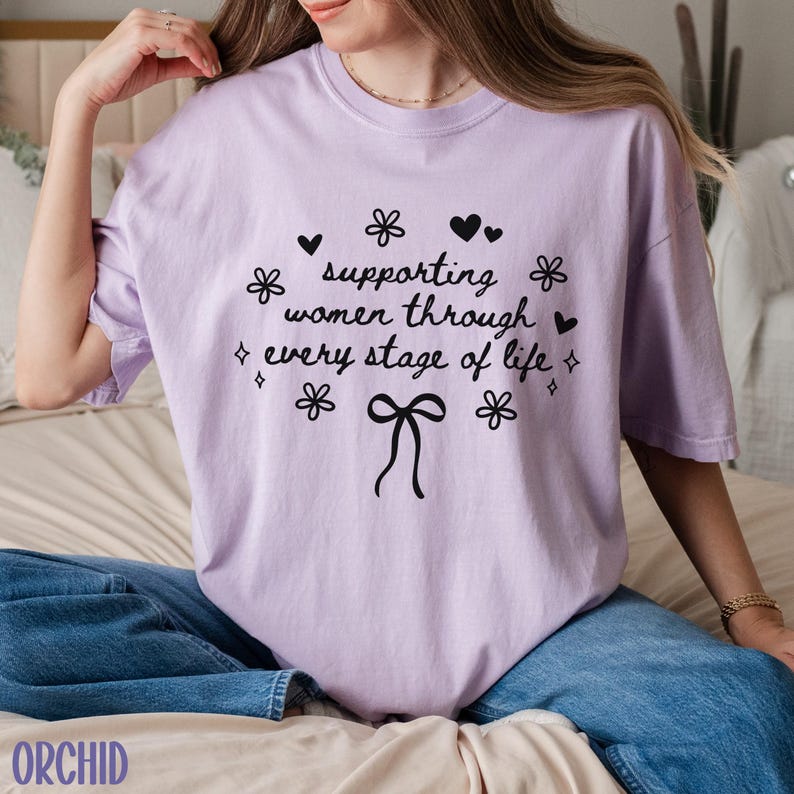 May include: A lavender t-shirt with a black graphic that reads "supporting women through every stage of life" with a bow and flowers.