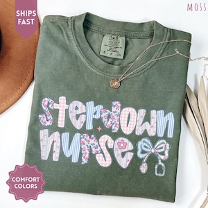 May include: An olive green Comfort Colors t-shirt with the words "Stepdown Nurse" in a colorful, patterned design. The design includes floral, gingham, and striped patterns, along with a bow and stethoscope graphic. A gold necklace is draped across the shirt.