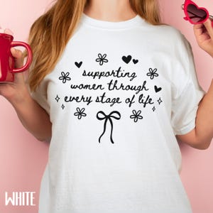 May include: White t-shirt with a black graphic that says "supporting women through every stage of life" with a bow and flowers.