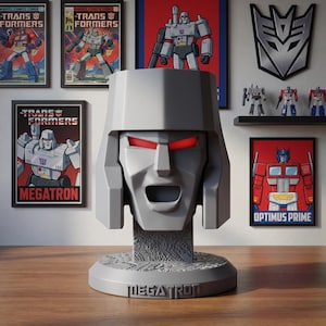 May include: A gray Megatron bust sculpture featuring a stylized robotic head with glowing red eyes and an open mouth. The base of the bust is circular with intricate mechanical detailing and the name Megatron embossed on the front surface.