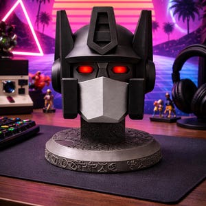 May include: A black and silver robotic head bust featuring glowing red eyes and a metallic faceplate. The figure sits on a circular base decorated with intricate, etched symbols, showcasing a detailed mechanical design with sharp, angular features.
