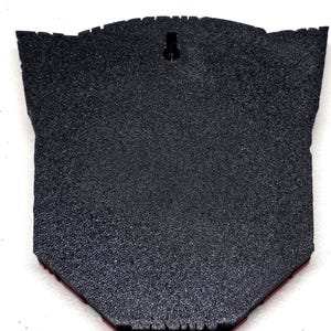 May include: A black, textured wall hanging with a unique shape. The back features a keyhole for hanging and a red border along the bottom edge. The surface has a speckled appearance.