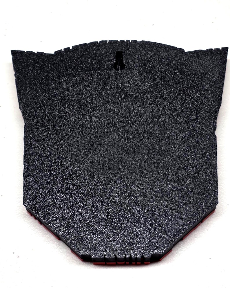 May include: A black, textured wall hanging with a unique shape. The back features a keyhole for hanging and a red border along the bottom edge. The surface has a speckled appearance.