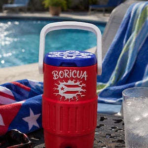 May include: Red and blue insulated water cooler with a white handle. The cooler has the word "BORICUA" and a graphic of the Puerto Rican flag. A glass of water and sunglasses sit nearby, with a pool in the background.