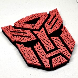 May include: A red and black emblem featuring the word "AUTOBOTS" and other character names. The emblem is a stylized shield shape, with the names of characters like Optimus Prime and Bumblebee.