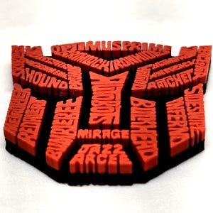May include: A red and black 3D-printed Transformers logo. The logo is filled with the names of various Transformers characters. The logo is red with a black border and sits on a white surface.