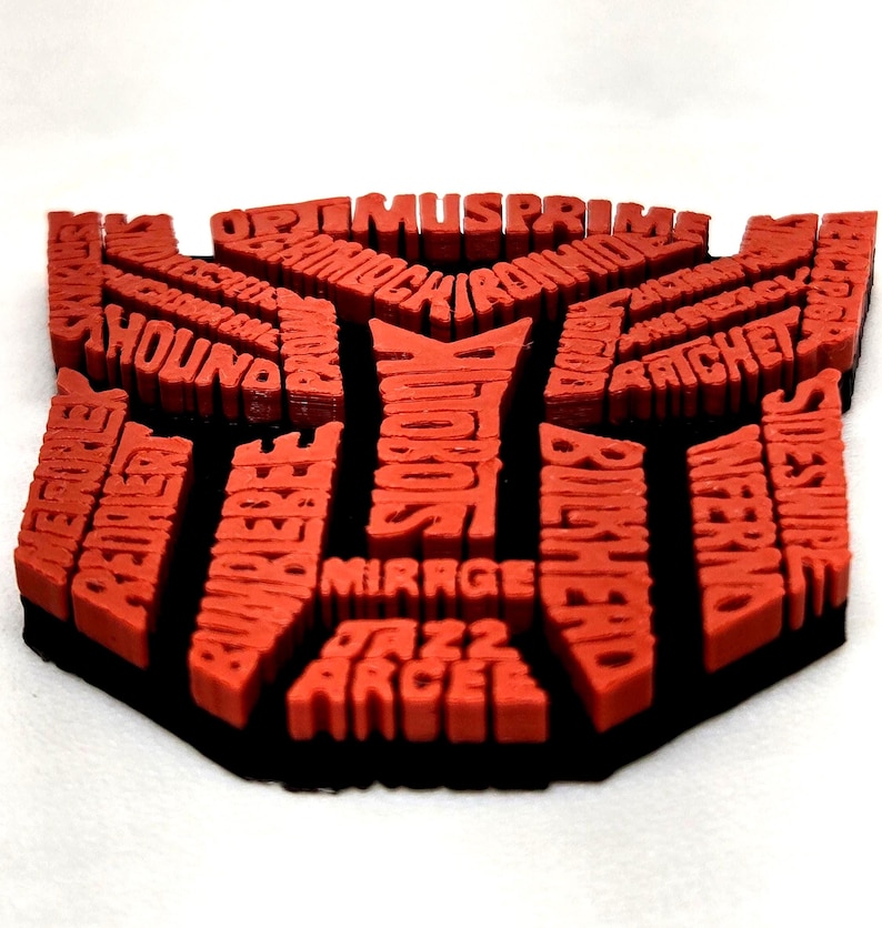 May include: A red and black 3D-printed Transformers logo. The logo is filled with the names of various Transformers characters. The logo is red with a black border and sits on a white surface.