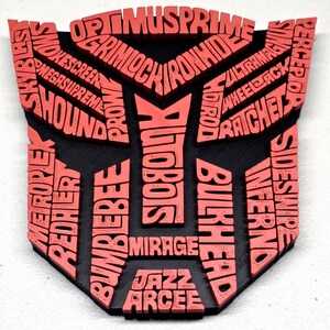 May include: A black and red emblem featuring the names of Transformers characters. The red text includes names like Optimus Prime, Bumblebee, and Autobots, set against a black background. The design is a stylized shield shape.