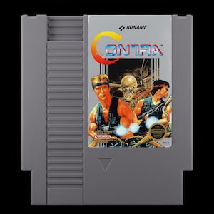 May include: A gray Nintendo Entertainment System (NES) cartridge for the game Contra. The label features two muscular men with guns, a menacing alien, and the game's title in a stylized font. The Konami and Nintendo logos are also visible.
