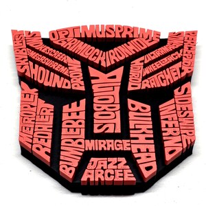 May include: A black and coral-colored Transformers logo featuring character names like Optimus Prime, Bumblebee, and Megasupreme. The logo is a shield shape with the names of the characters in a 3D effect.
