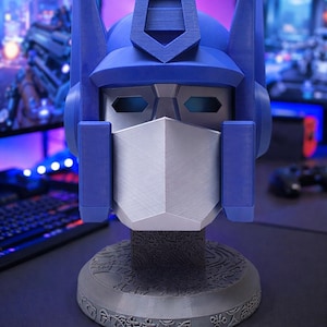 May include: A blue and silver Optimus Prime bust, a collectible item. The bust features a detailed design of the character's head, with a silver faceplate and blue helmet. The base is gray with engraved symbols. A gaming setup is in the background.
