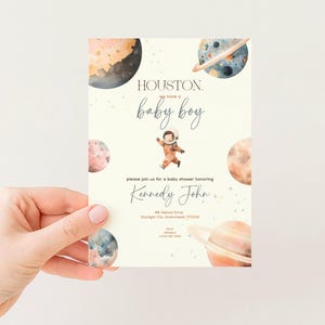 May include: A baby shower invitation with a space theme. The invitation features watercolor planets, an astronaut illustration, and the text "Houston, we have a baby boy." The invitation is for Kennedy John.