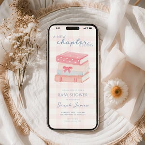 May include: A smartphone displays a baby shower invitation with a stack of watercolor books. The text reads "A New Chapter Begins" and invites guests to celebrate Sarah James. The background is a soft, pink gingham pattern.